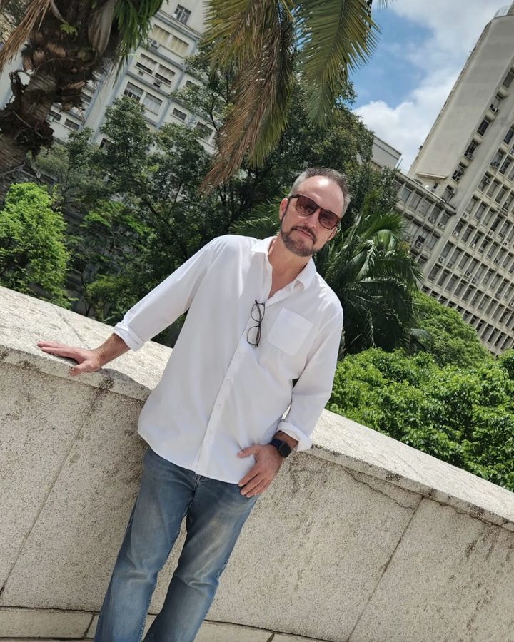 Vaughn Bullard in a white shirt and jeans, leaning on a curved stone wall among palm trees