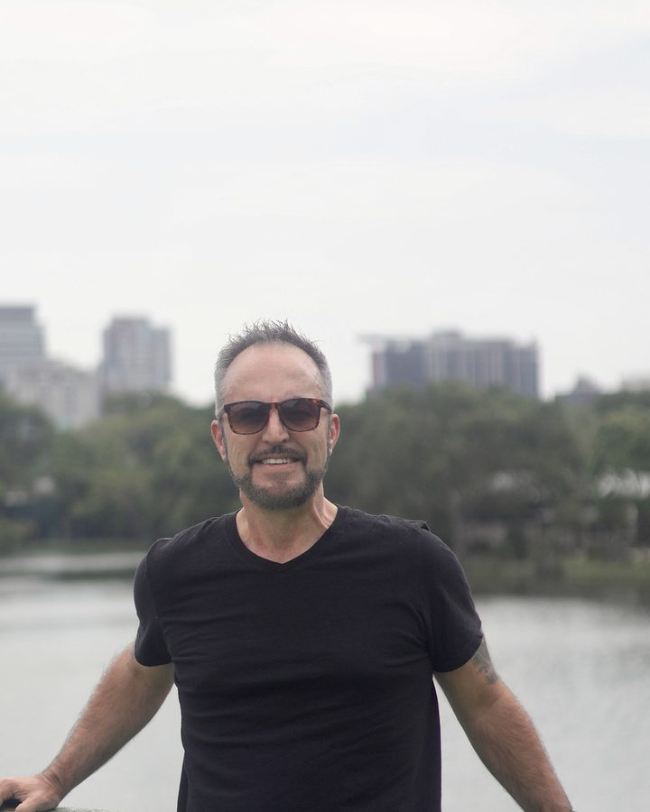 Vaughn Bullard smiling in a black T-shirt and sunglasses, standing at a green railing by the water