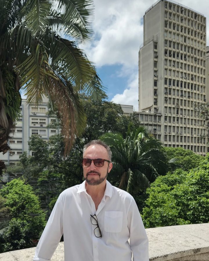 Vaughn Bullard in a white shirt and jeans, standing against a stone wall with trees and tall buildings behind
